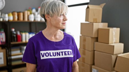 Mature woman volunteer in purple shirt standing in a warehouse with cardboard boxes.