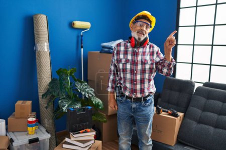 Photo for Cheerful senior man, safety glasses and hardhat on, points sideways in his fresh new home! looking confident, handyman's happy welcome gesture captured! - Royalty Free Image