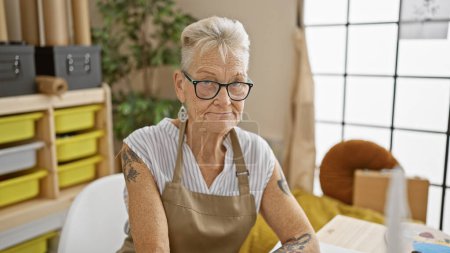 Téléchargez les photos : Femme artiste senior aux cheveux gris, avec un visage sérieux, peint intensément alors qu'elle est assise à une table dans son atelier - en image libre de droit