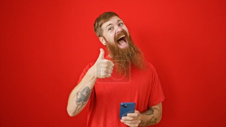 Téléchargez les photos : Joyeux, confiant jeune roux homme clignote un pouce vers le haut tout en envoyant un message joyeux sur son smartphone, le tout sur un fond rouge isolé vibrant - en image libre de droit