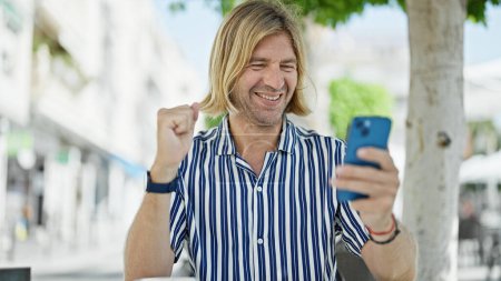Téléchargez les photos : Un homme blond joyeux célèbre en utilisant un smartphone sur une terrasse de la rue de la ville. - en image libre de droit