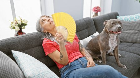 In the heat at home, a mature middle age, grey-haired woman sitting comfortably on the cozy sofa, relaxes fanning air with a hand fan, while her beloved pet dog sits beside her
