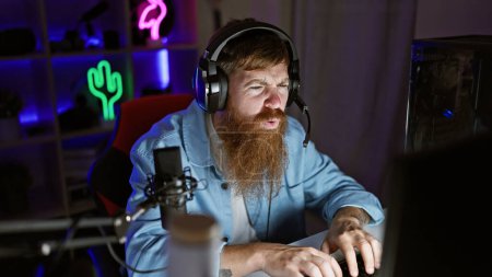 Schöner irischer Rotschopf Streamer in ernstem Spiel auf seinem geschmückten Gaming-Rig gesperrt, Nacht-Streaming-Sitzung aus seinem geschmückten Spielzimmer