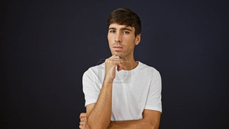 Serious yet relaxed, a cool young hispanic man with a doubtful expression stands isolated against a black background. pondering, mind full of ideas, this attractive male gives a captivating portrait.