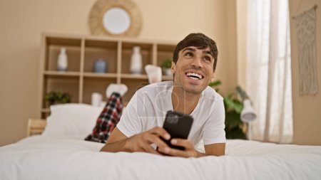 Foto de Atractivo, joven hispano disfrutando de una mañana relajada, cómodamente acostado en la cama, felizmente mensajes de texto en el teléfono inteligente con una sonrisa reluciente en un acogedor dormitorio interior de la casa - Imagen libre de derechos