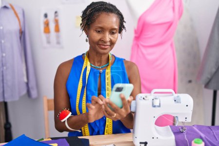 Téléchargez les photos : Moyen Age femme afro-américaine tailleur souriant confiant en utilisant smartphone à l'atelier - en image libre de droit