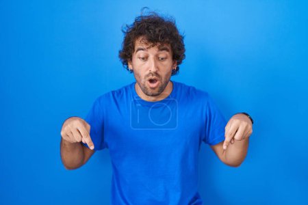 Téléchargez les photos : Jeune homme hispanique debout sur fond bleu pointant vers le bas avec les doigts montrant la publicité, visage surpris et bouche ouverte - en image libre de droit