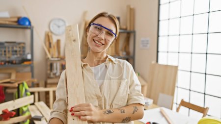 Téléchargez les photos : Une femme souriante avec des lunettes de sécurité tient du bois dans un atelier de menuiserie lumineux. - en image libre de droit