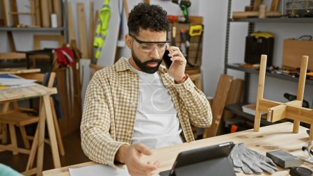 Photo for Young hispanic man with beard multitasking on a phone call in a carpentry workshop. - Royalty Free Image