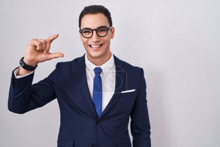 Téléchargez les photos : Jeune homme hispanique portant costume et cravate geste souriant et confiant avec la main faisant signe de petite taille avec les doigts à la recherche et la caméra. concept de mesure. - en image libre de droit