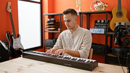 Photo for Handsome hispanic man playing keyboard in a modern music studio with guitars and podcasting equipment. - Royalty Free Image