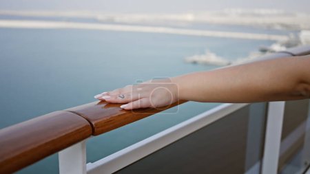 A close-up view of a woman's hand resting on the railing of a cruise ship deck with the ocean horizon in the background.