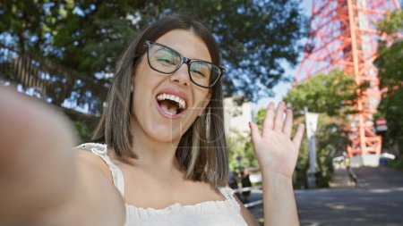 Selbstbewusste, schöne hispanische Frau genießt Tokyo, winkt Hallo, während sie Spaß macht, lässiges Selfie mit aufgesetzter Brille, Ausdruck von Glück im Straßenporträt der Stadt.