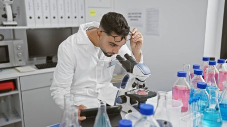 Foto de Un hombre en un laboratorio examina muestras a través de un microscopio rodeado de tubos de ensayo, transmitiendo investigación y ciencia. - Imagen libre de derechos