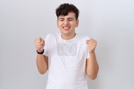 Téléchargez les photos : Jeune homme non binaire portant t-shirt blanc décontracté excité pour le succès avec les bras levés et les yeux fermés célébrant la victoire en souriant. concept gagnant. - en image libre de droit