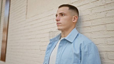 Hombre hispano joven guapo en ropa casual posando con una expresión reflexiva sobre un fondo de pared de ladrillo urbano.