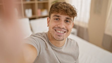 Joven hispano haciendo videollamada sentado en la cama en el dormitorio