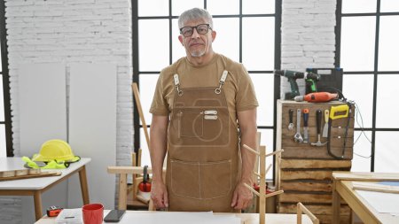 Téléchargez les photos : Homme mûr dans tablier se tient avec confiance dans un atelier de menuiserie bien équipé avec des outils et des meubles en bois autour. - en image libre de droit