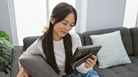 A young asian woman sitting on a couch at home, smiling while browsing on a tablet indoors.