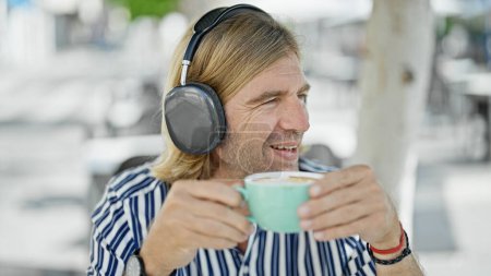 Téléchargez les photos : Un homme souriant dégustant un café à l'extérieur tout en écoutant de la musique avec des écouteurs dans un cadre urbain. - en image libre de droit