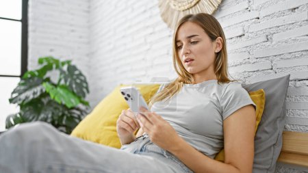 Téléchargez les photos : Jeune femme blonde séduisante assise confortablement sur son lit, immergée dans l'utilisation de son smartphone dans une matinée détendue dans la chambre confortable de sa maison - en image libre de droit