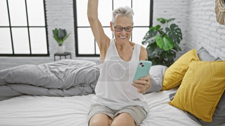 Téléchargez les photos : Vue chaleureuse d'une femme âgée souriante aux cheveux gris célébrant une victoire, assise confortablement sur son lit en pyjama, envoyant des messages matinaux à l'aide de son smartphone à la maison dans sa chambre confortable - en image libre de droit