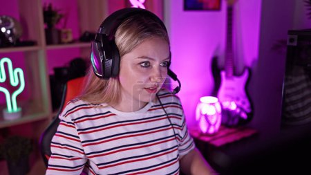 Photo for A young blonde woman wearing headphones in a colorful gaming room looking focused and engaged. - Royalty Free Image