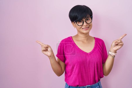 Téléchargez les photos : Jeune femme asiatique aux cheveux courts debout sur fond rose souriant confiant pointant avec les doigts vers différentes directions. espace de copie pour la publicité - en image libre de droit