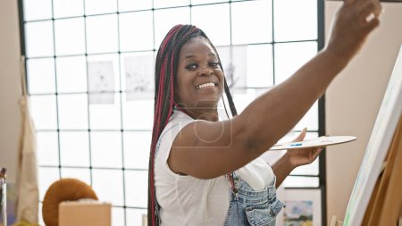 Joyful african american female artist draws confidently, smiling radiantly in her art studio while standing over her canvas, embracing her creativity