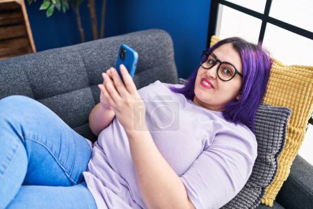 Foto de Joven hermosa mujer de tamaño grande utilizando el teléfono inteligente acostado en el sofá en casa - Imagen libre de derechos