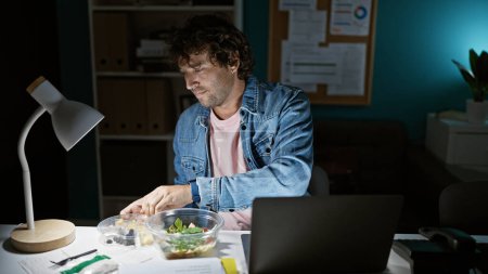A young hispanic man eating salad while working late in the office, illuminated by a desk lamp.