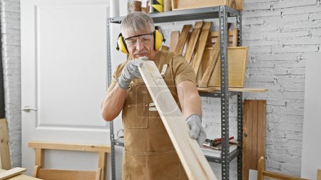 Téléchargez les photos : Un homme mûr inspecte le bois dans son atelier de menuiserie tout en portant un équipement de protection. - en image libre de droit