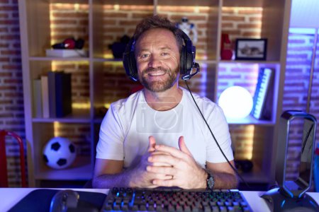 Photo for Middle age man with beard playing video games wearing headphones with hands together and crossed fingers smiling relaxed and cheerful. success and optimistic - Royalty Free Image