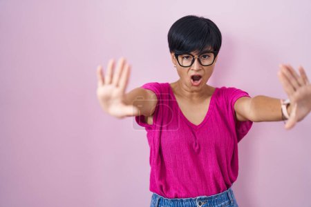 Foto de Joven mujer asiática con pelo corto de pie sobre fondo rosa haciendo stop gesture con palmas de las manos, enojado y expresión de frustración - Imagen libre de derechos