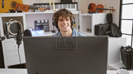 Photo for A joyful young man with headphones working in a music studio full of instruments and recording equipment. - Royalty Free Image