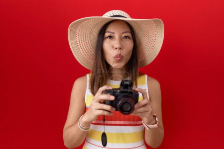 Photo for Middle age chinese woman using reflex camera wearing summer hat looking at the camera blowing a kiss being lovely and sexy. love expression. - Royalty Free Image