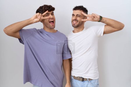Foto de Homosexual pareja gay de pie sobre blanco fondo haciendo la paz símbolo con los dedos sobre la cara, sonriendo alegre mostrando victoria - Imagen libre de derechos
