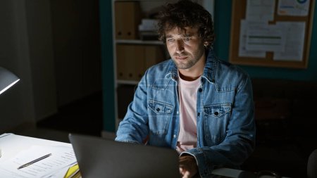 Foto de Hombre hispano guapo trabajando hasta tarde en la oficina, rodeado de documentos y una computadora portátil. - Imagen libre de derechos