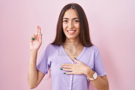 Foto de Mujer hispana joven con el pelo largo de pie sobre fondo rosa sonriendo jurando con la mano en el pecho y los dedos hacia arriba, haciendo una promesa de lealtad juramento - Imagen libre de derechos