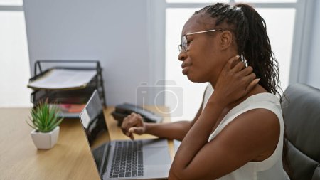 Foto de Mujer afroamericana que experimenta dolor de cuello mientras trabaja en una computadora portátil en un entorno de oficina moderno. - Imagen libre de derechos