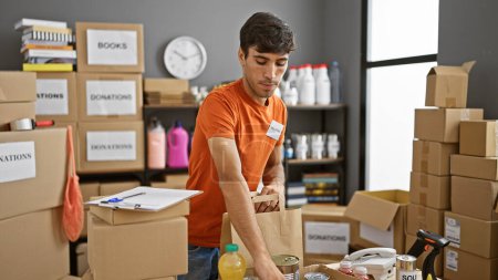Photo for Handsome young hispanic man actively volunteering at local charity center, reading clipboard, checking donated products, packaging food in cardboard boxes, portrait of altruism in action - Royalty Free Image