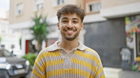 Selbstbewusster junger arabischer Mann, der fröhlich seine Zeit im Freien genießt, auf der Straße der Stadt stehend, sein bärtiges Gesicht, das durch ein hübsches Lächeln pure Freude und Glück ausdrückt