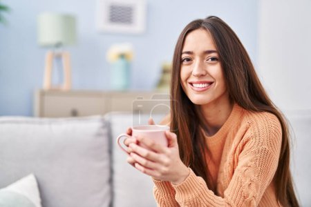 Foto de Joven mujer hispana hermosa bebiendo café sentado en el sofá en casa - Imagen libre de derechos