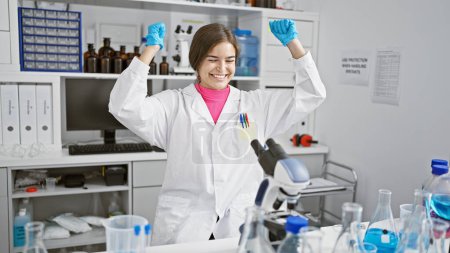 Foto de Joven hermosa mujer hispana científica usando microscopio celebrando en laboratorio - Imagen libre de derechos