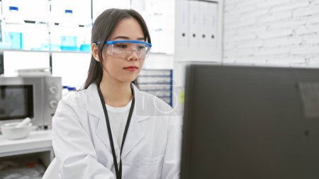 A focused asian woman scientist in a laboratory, wearing a white lab coat, safety glasses, analyzing data on a computer.