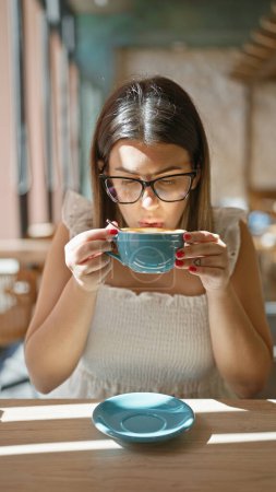 Beautiful hispanic woman enjoying her hot coffee drink inside sunny cafe, relaxing at breakfast while wearing glasses, sitting casually at cafeteria table enjoying the morning sun.