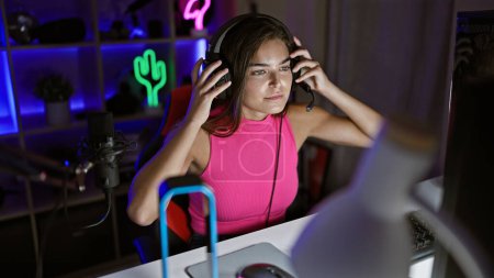 Téléchargez les photos : Attrayant jeune hispanique streamer féminin, pris dans la cyber nuit, entièrement concentré et portant des écouteurs tout en jouant à un jeu vidéo à enjeux élevés dans sa salle de jeu sombre à la maison - en image libre de droit