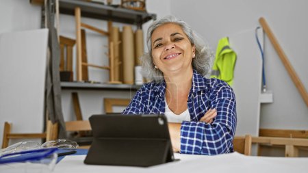 Photo for A smiling grey-haired woman in a checkered shirt sits confidently in a well-lit carpentry workshop. - Royalty Free Image