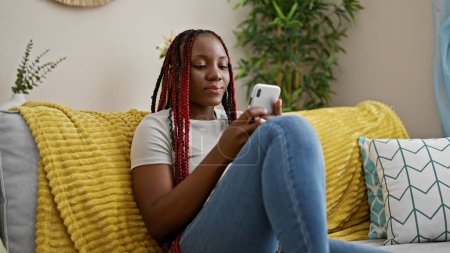 Photo for Beautiful african american woman, relaxed at home, intensely concentrates on typing a message on her phone as she sits on the sofa. - Royalty Free Image