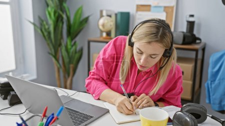 Photo for Blonde woman working indoors at office with laptop and headphones, taking notes while focused. - Royalty Free Image